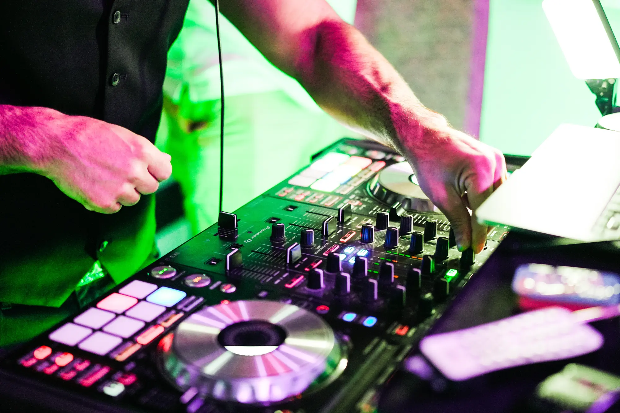 A DJ's hands on a controller with a jog wheel, faders and colored pads, lit in green and pink.