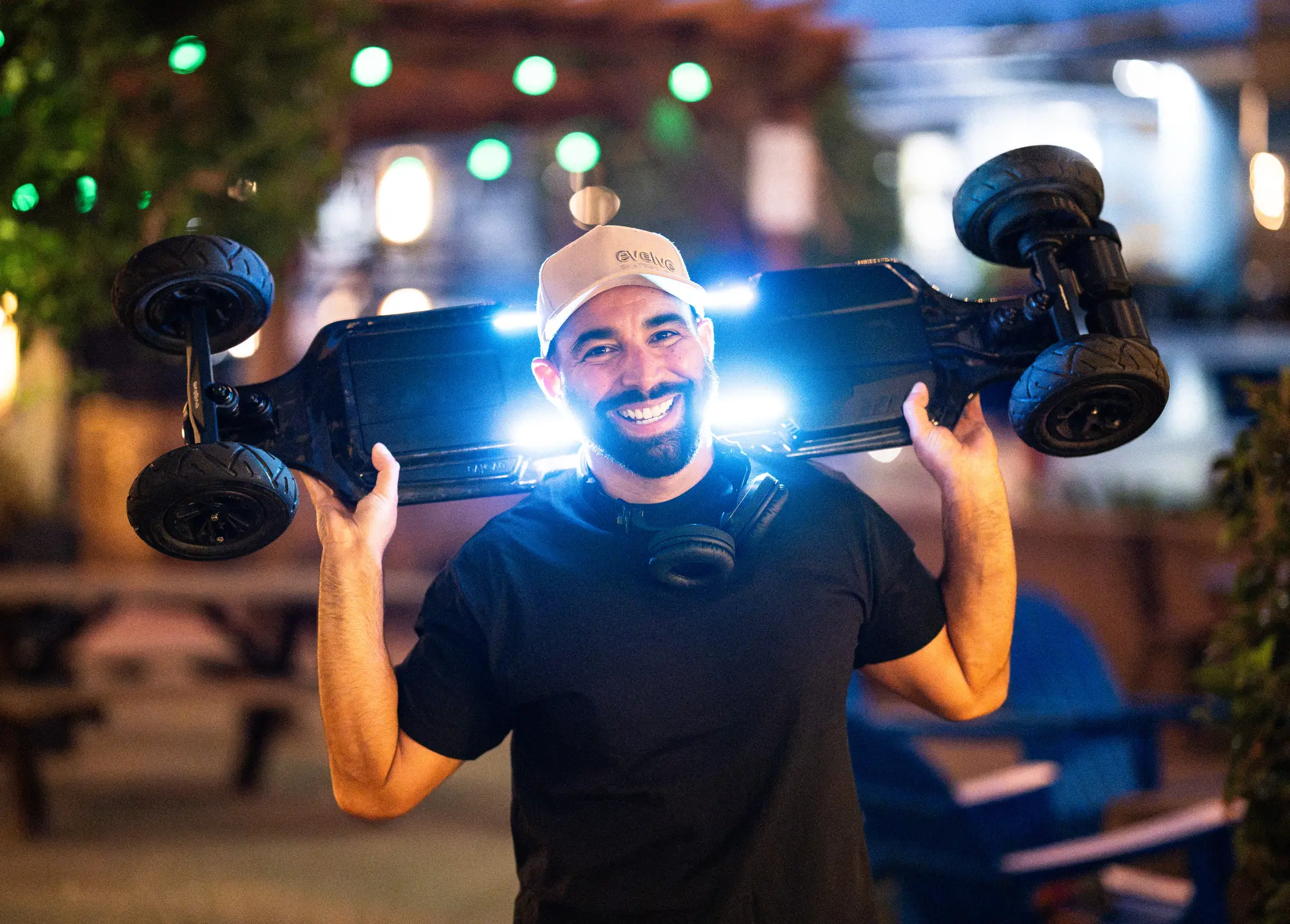 DJ Mojo smiling as he holds an electric skateboard over his head on an evening patio