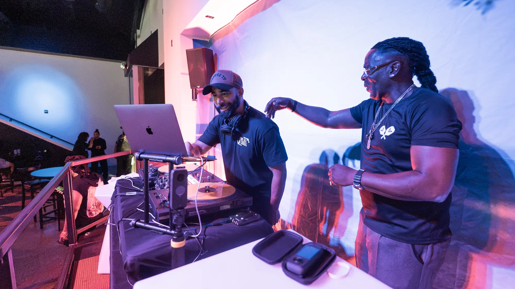 DJ Mojo in a cap and headphones at his laptop and turntable, with a smiling guest in a black t-shirt reaching toward the booth.