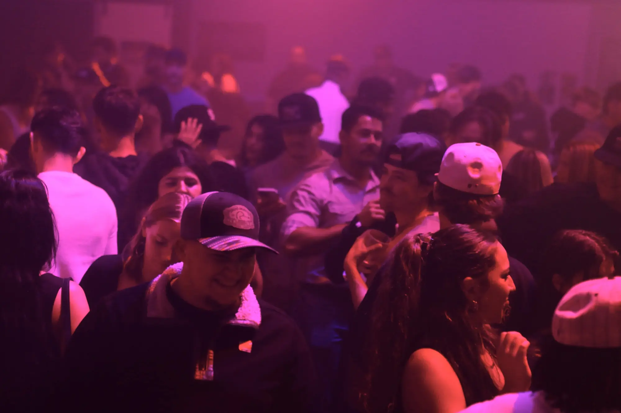 A crowded dance floor in pink haze with a smiling man in a cap in the middle of the guests.