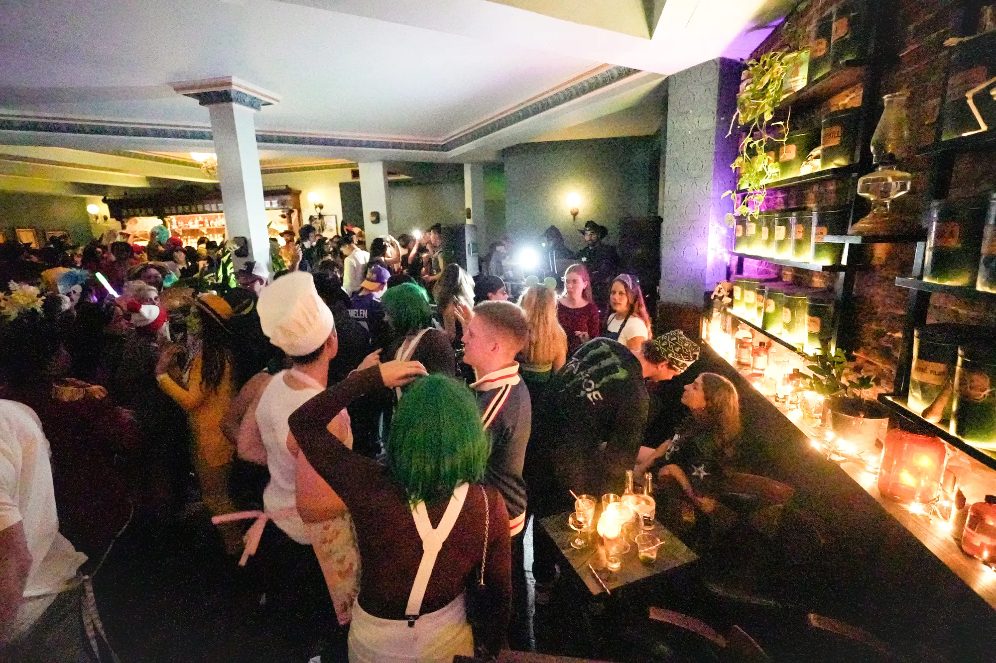 A packed room of costumed guests dancing in the glow of candles, with a woman in a green wig and a chef hat in the middle.