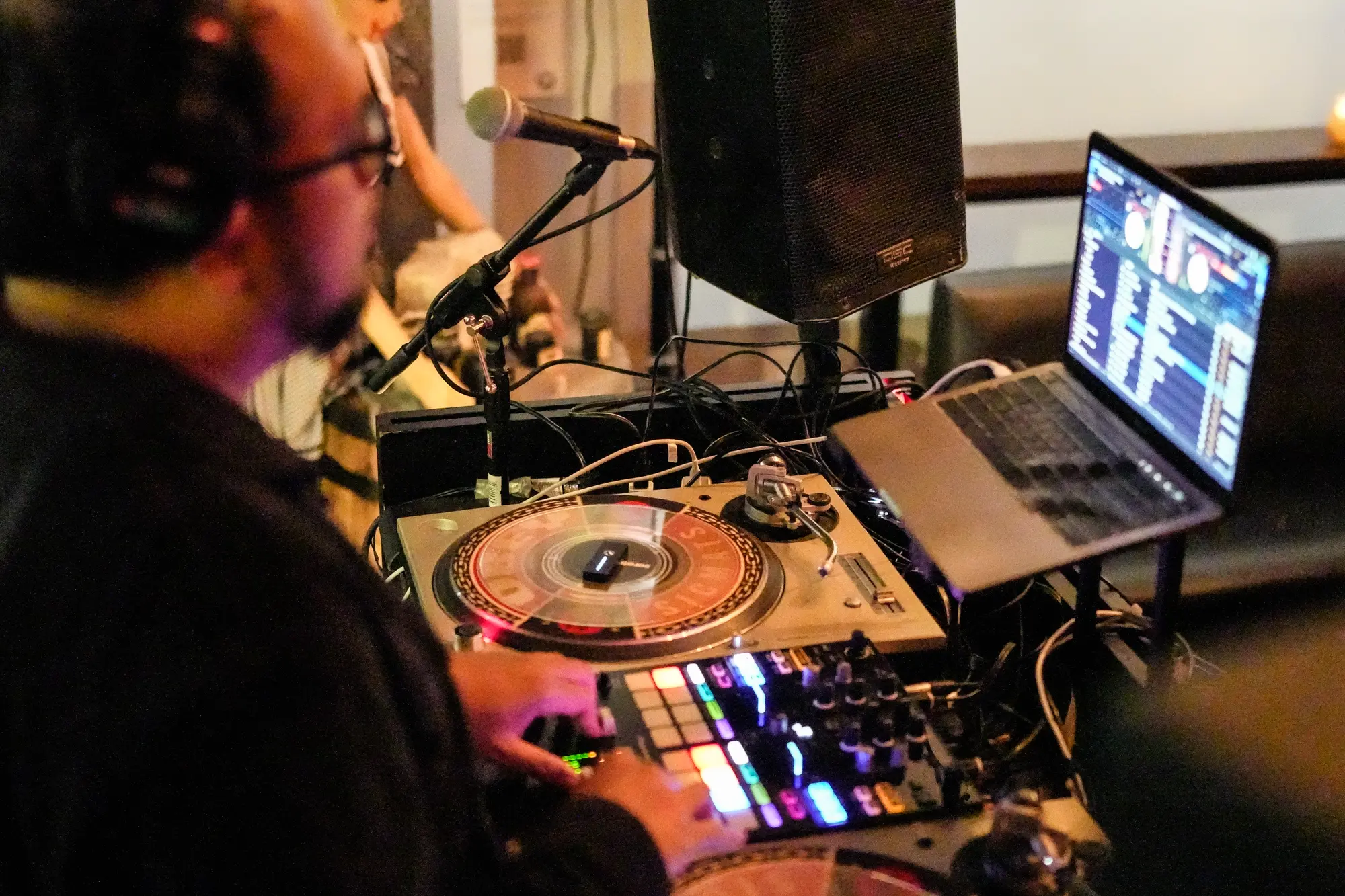 DJ Mojo at his turntables and laptop with a microphone and speaker beside the booth.
