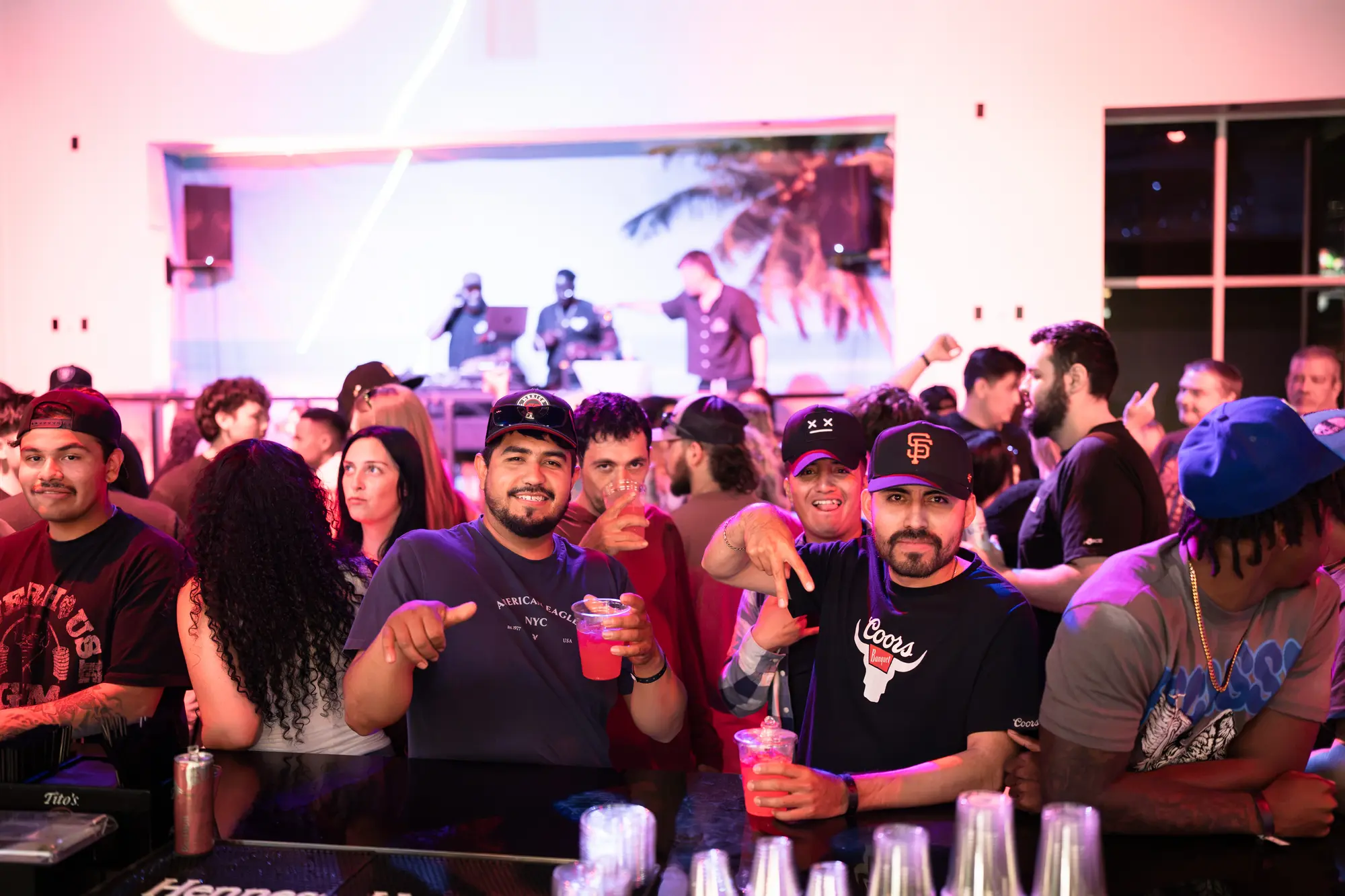 Guests at the bar cheer and point at the camera while DJs play on a stage behind them